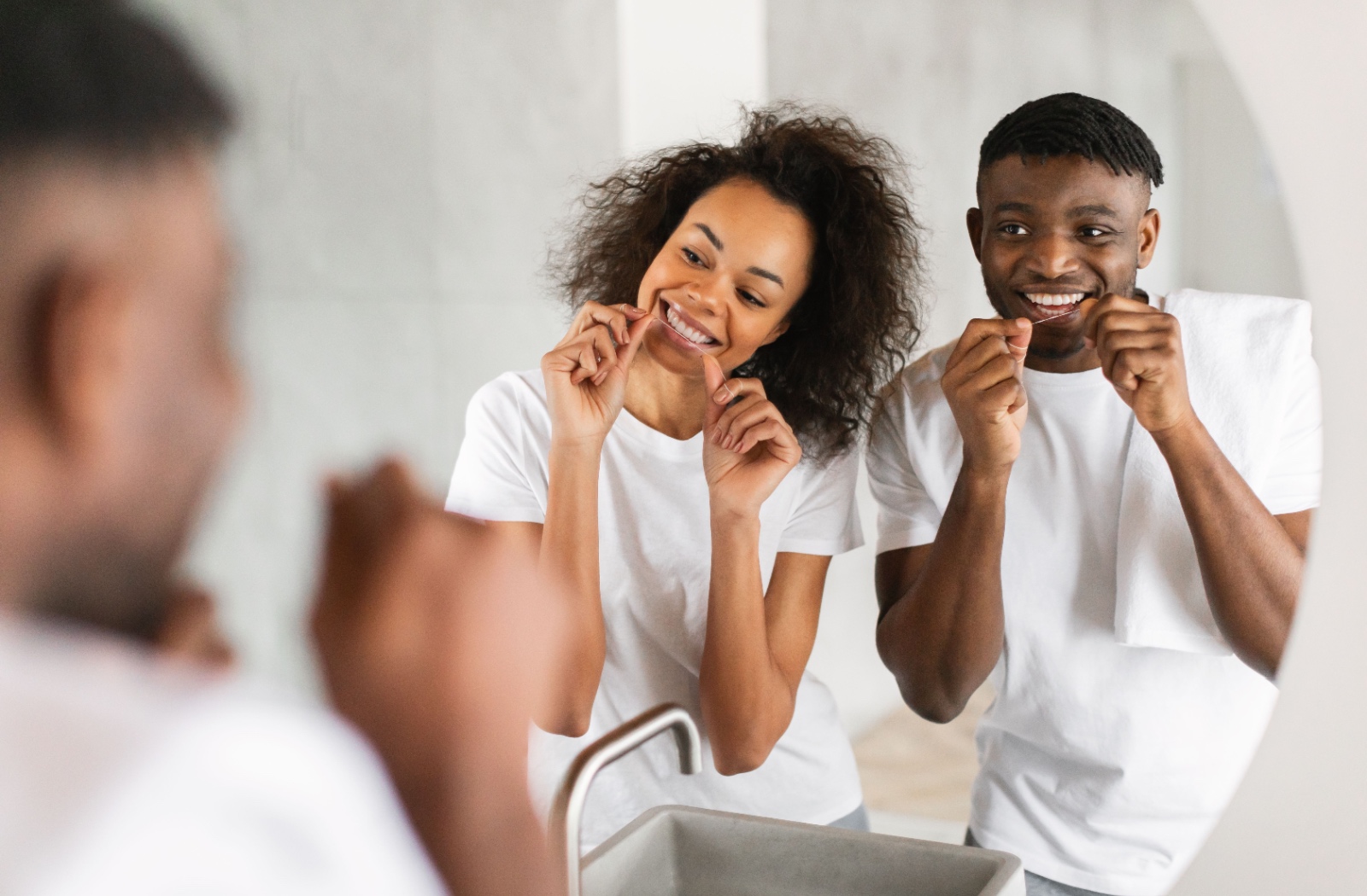 Smiling couple flossing their teeth together in front of a bathroom mirror.