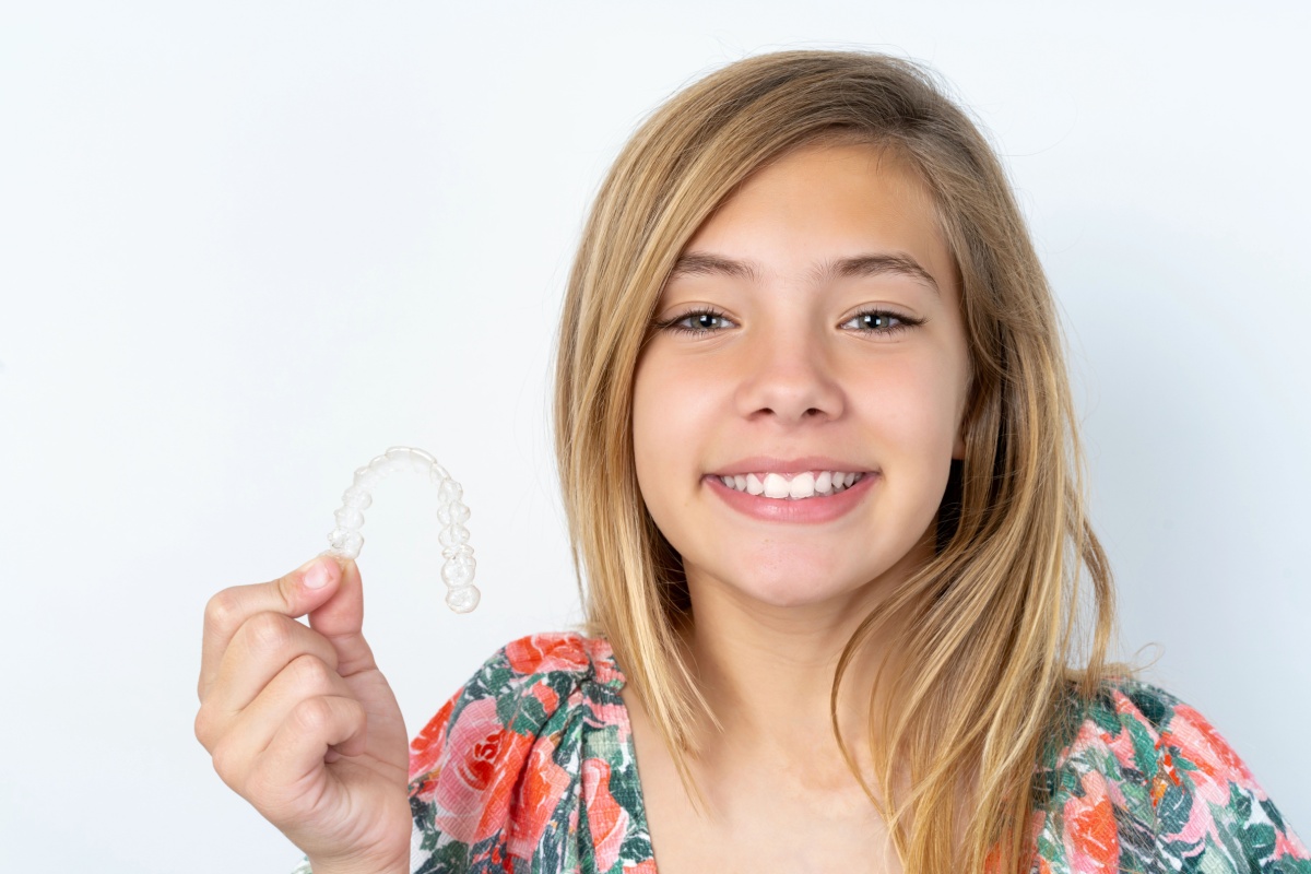 Adolescente souriante aux cheveux blonds tenant une gouttière d'alignement transparent, démontrant un traitement orthodontique Invisalign.